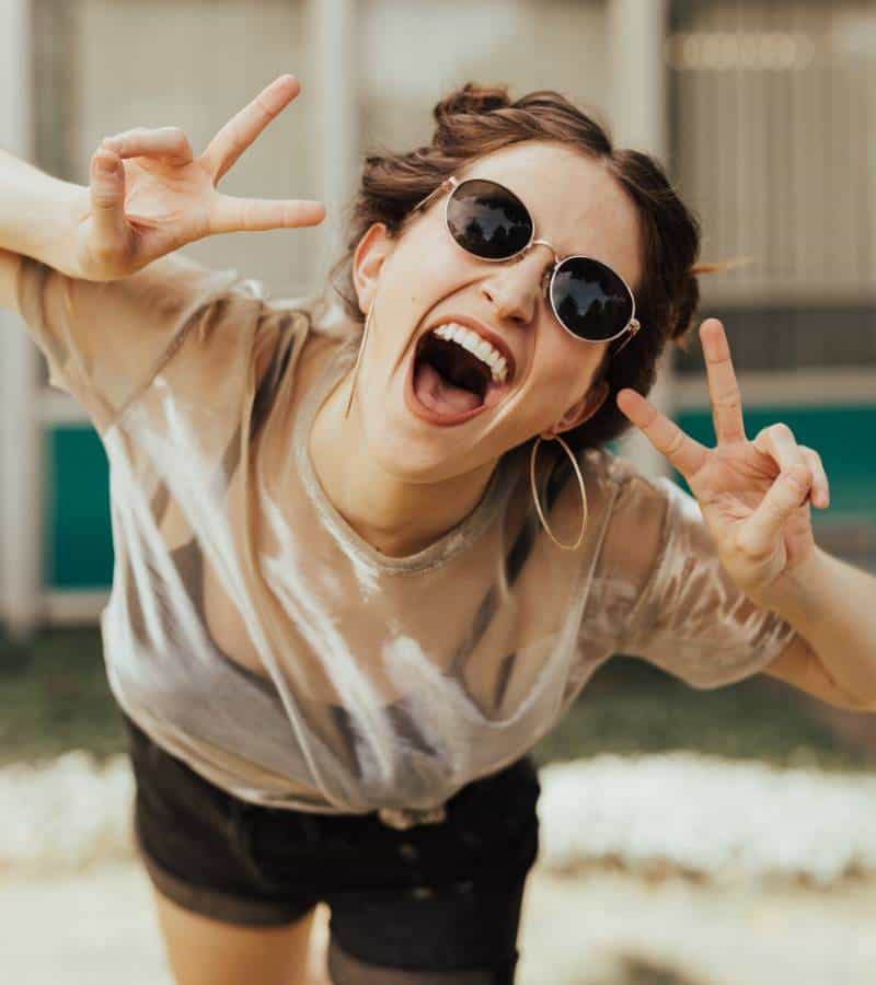 Happy woman in sunglasses and see-through top giving peace signs with fingers