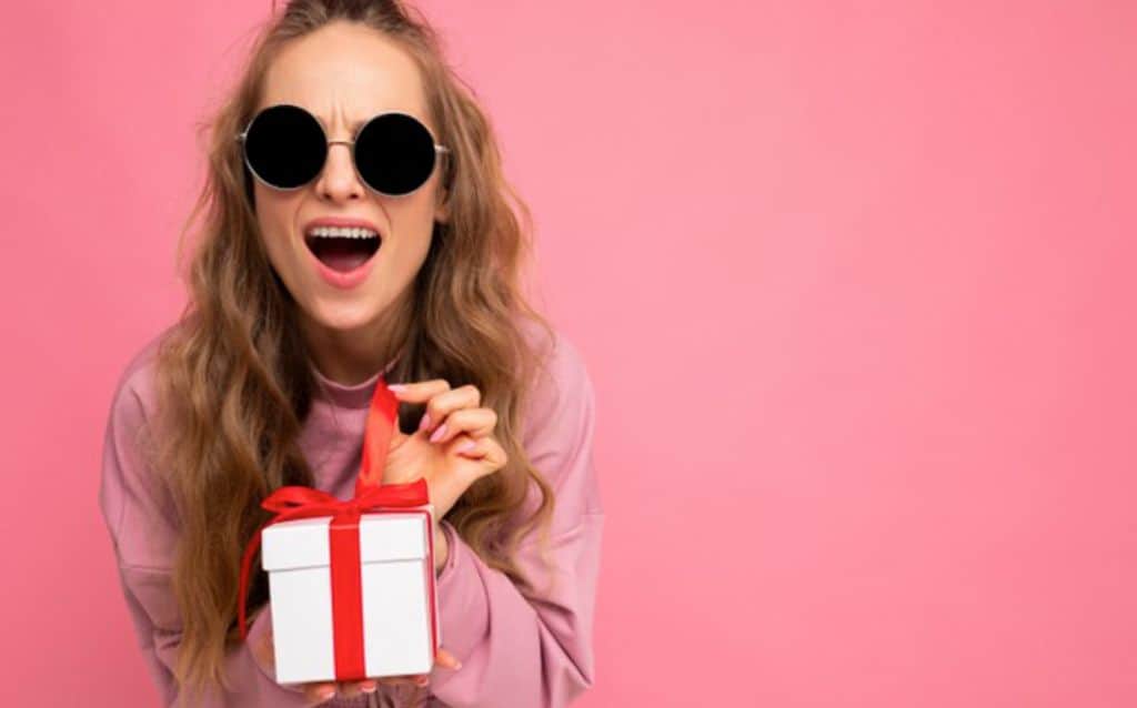 Girl in dark sunglasses pulling ribbon on present | Pink background photoshoot